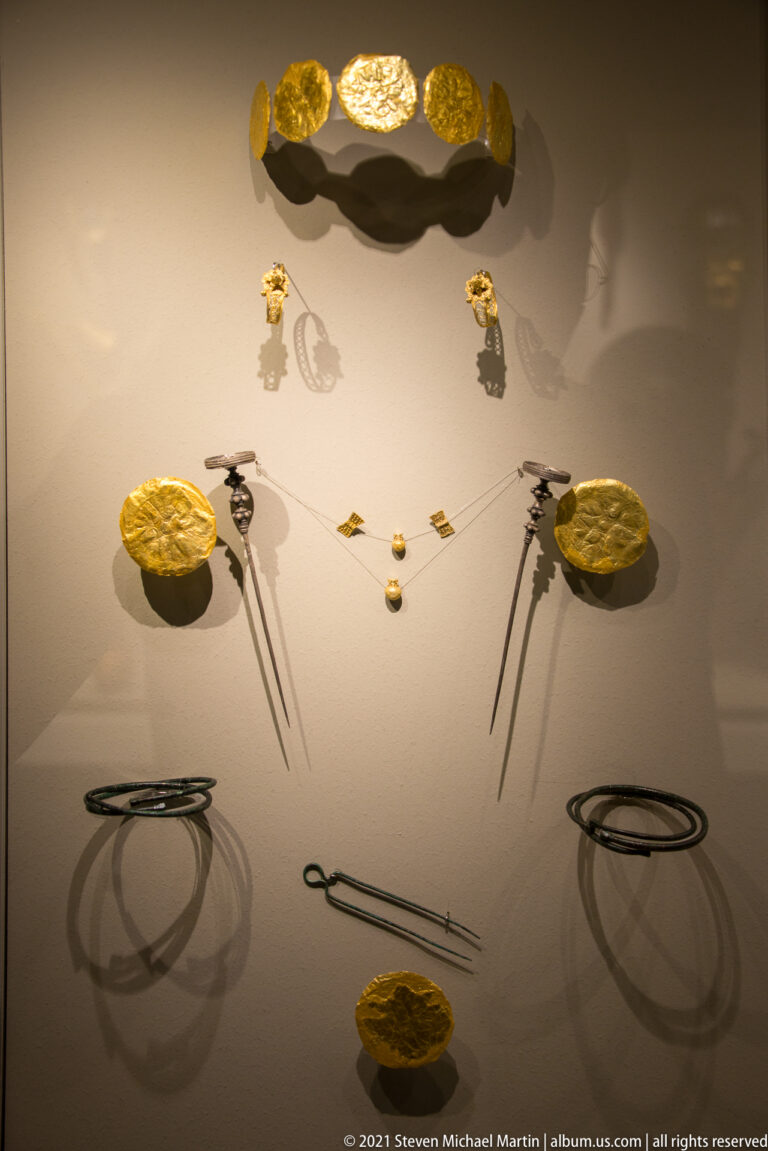Gold rosettes, long pins, small pendants, and dark metal bracelets arranged symmetrically in a museum case.