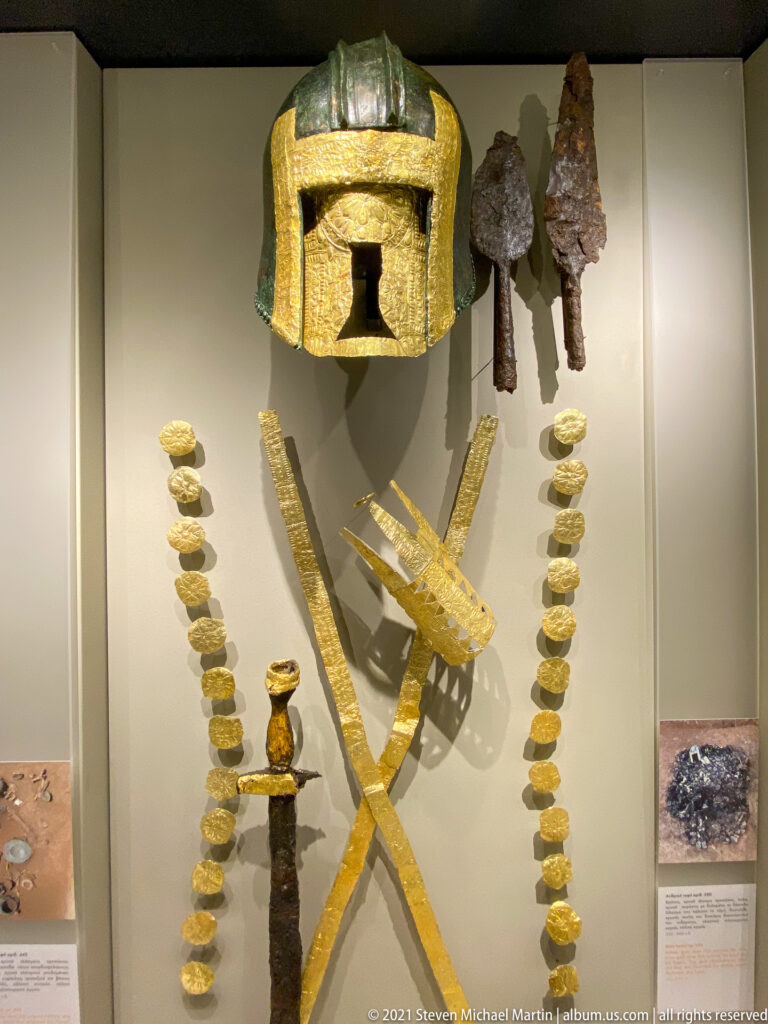 Museum case with bronze helmet, corroded weapons, crossed gold bands, and two vertical rows of small gold ornaments.