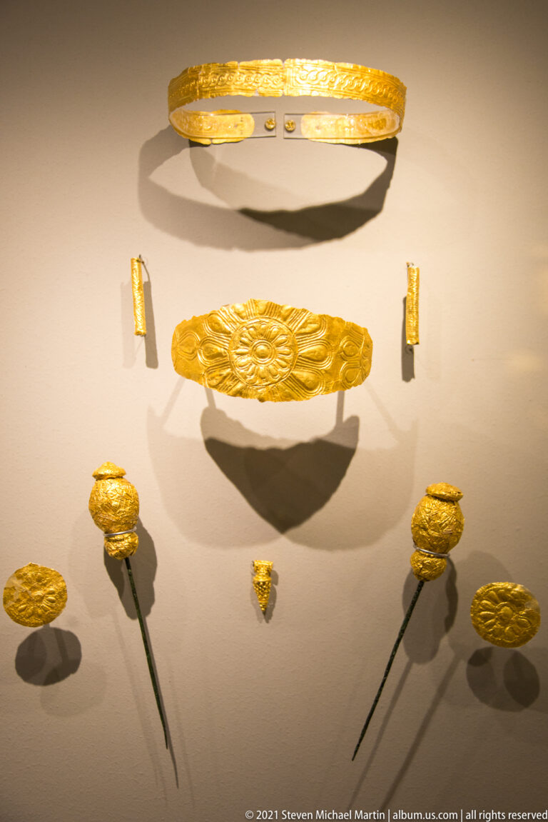 Embossed gold band, broad plaque, rosettes, small fittings, and two long pins mounted in a museum case.