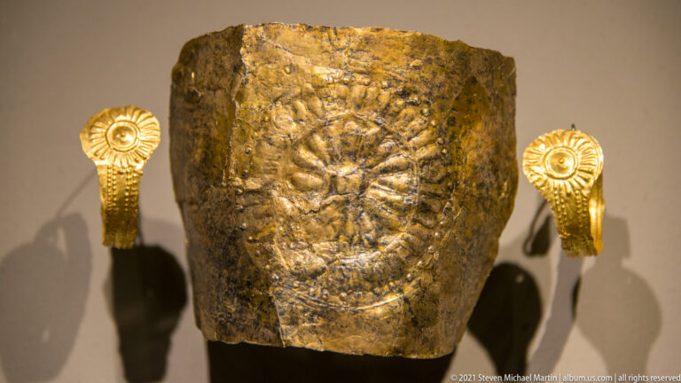 Curved embossed gold panel with a central face-like motif, flanked by two smaller spiral-ended gold fittings.