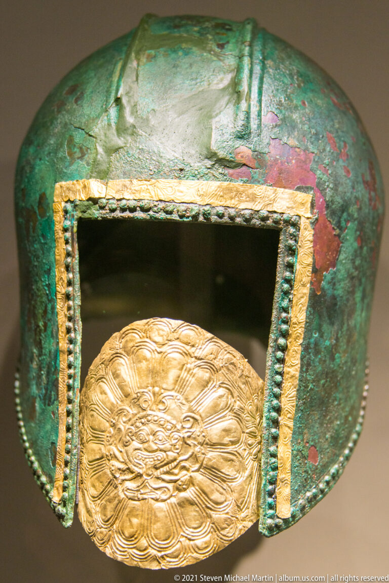 Green-patinated bronze helmet with gold-edged face opening and large embossed gold medallion across the lower front.