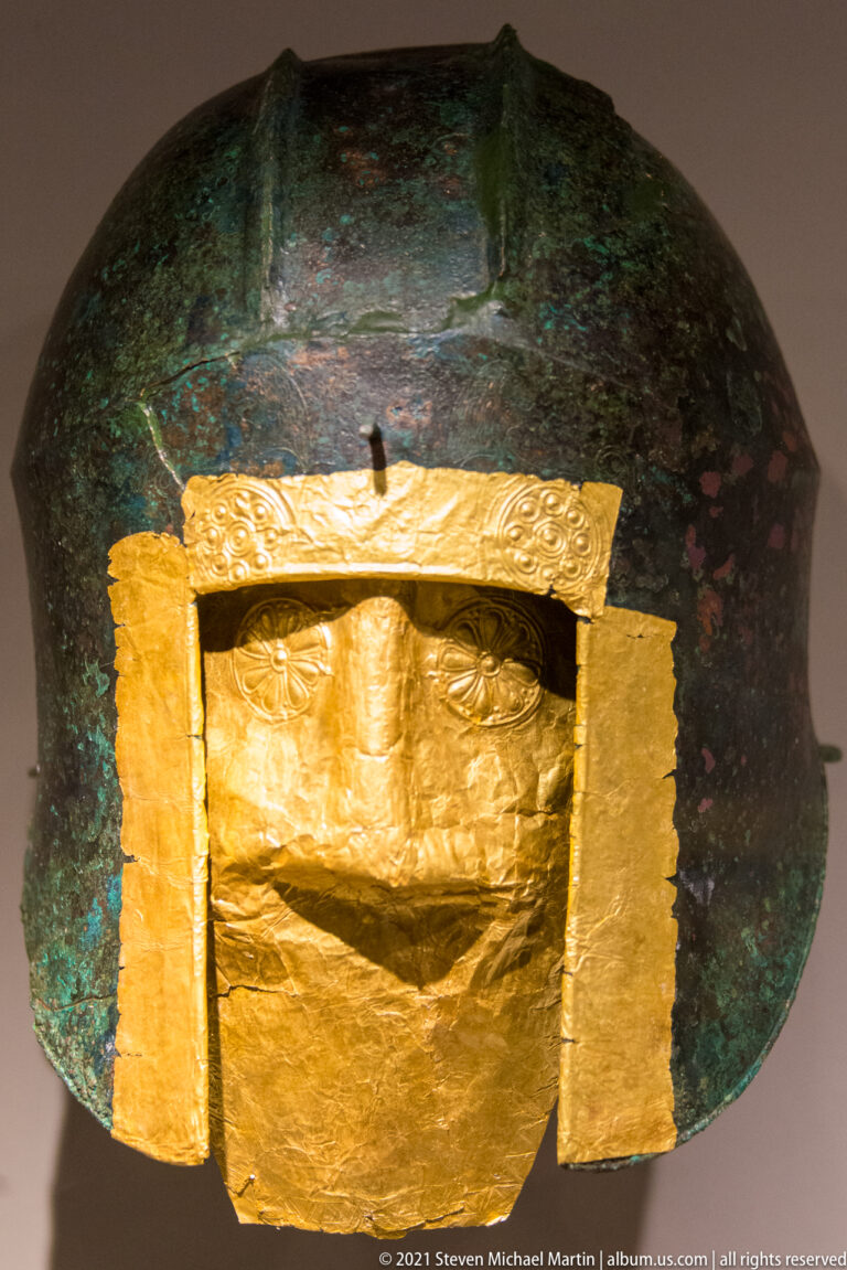 Bronze helmet with green patina and a large rectangular wrinkled gold sheet covering the front opening.