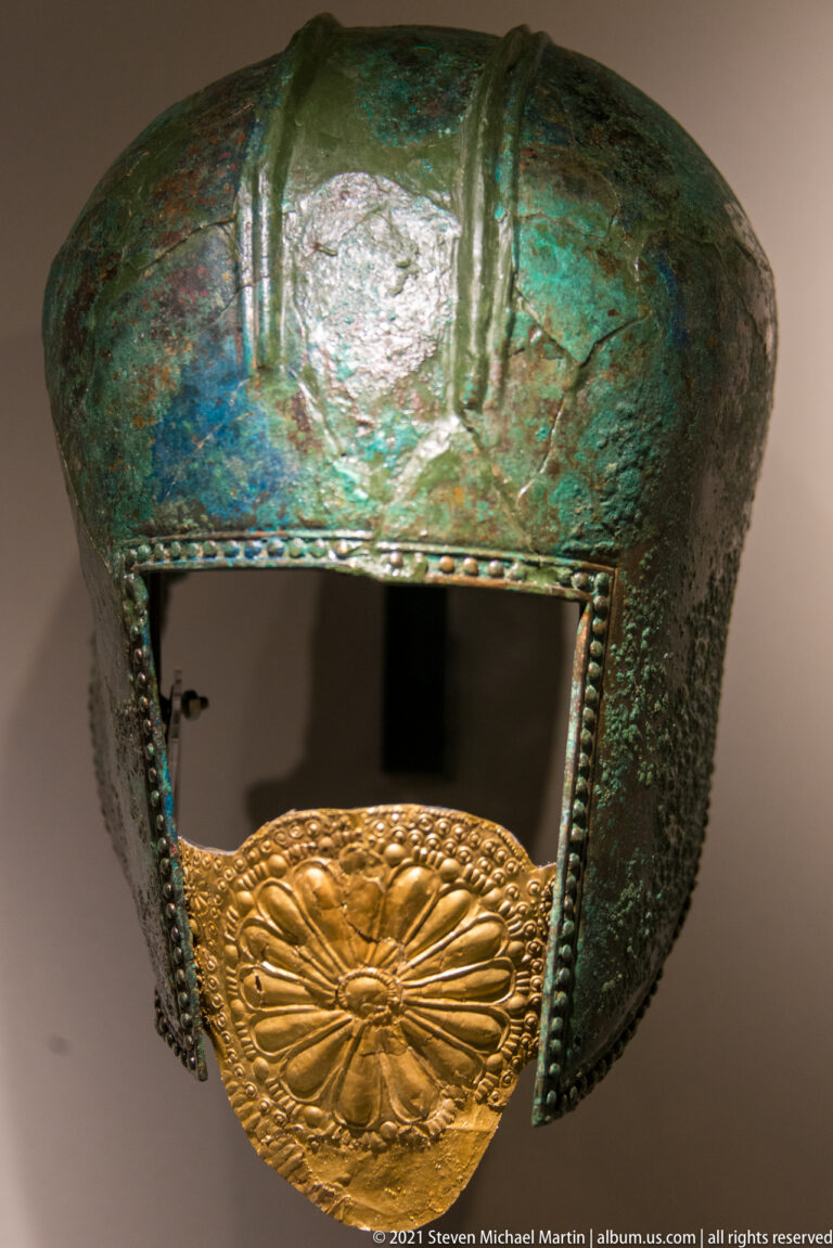 Green-patinated bronze helmet with rectangular face opening and a large embossed gold floral plaque below.