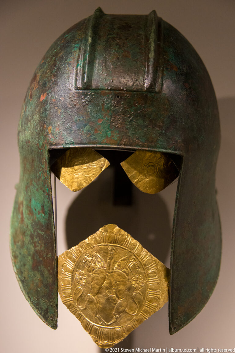 Bronze helmet with green patina and several embossed gold plaques attached around the lower front opening.