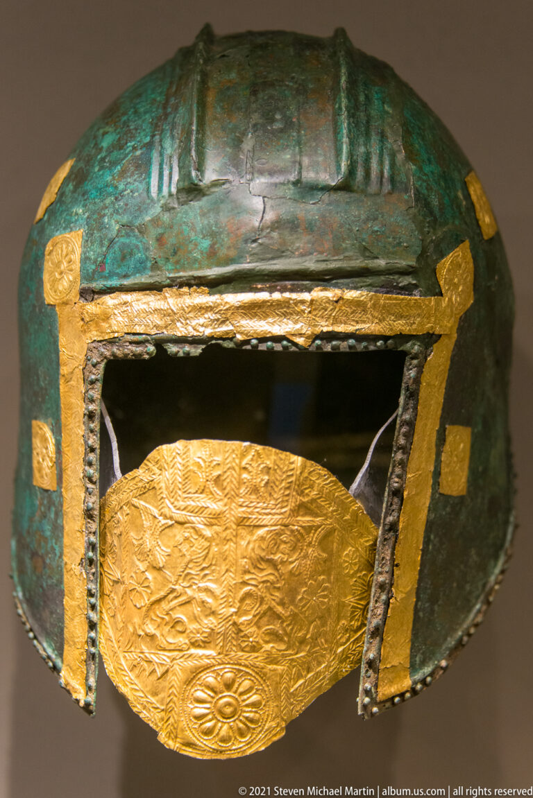 Bronze helmet with gold frame around the face opening and a large embossed oval gold plaque below.