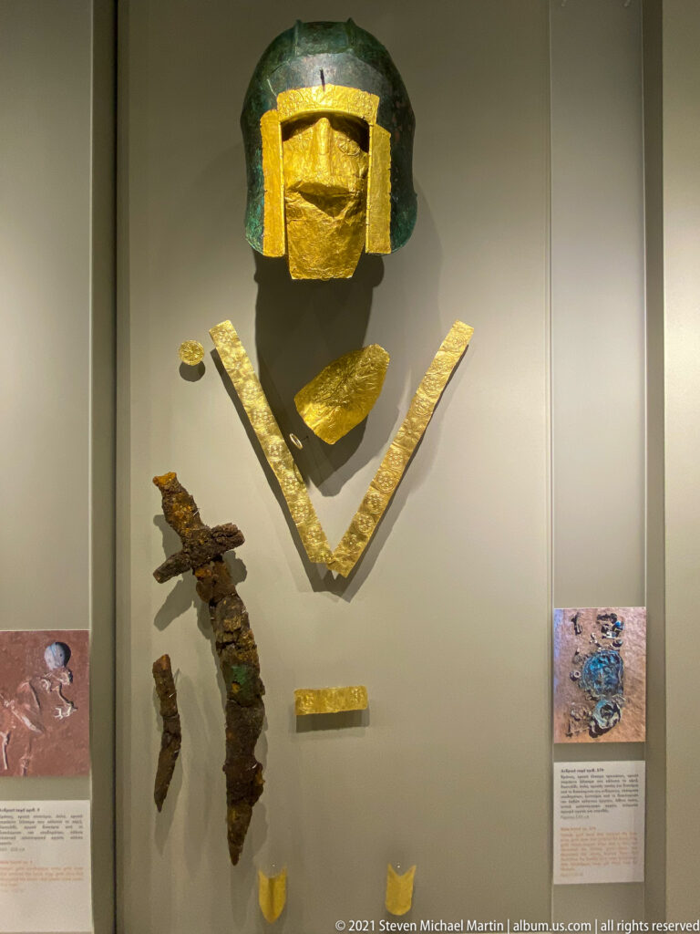 Bronze helmet with rectangular gold facing above angled gold bands and corroded iron weapon fragments in a museum case.