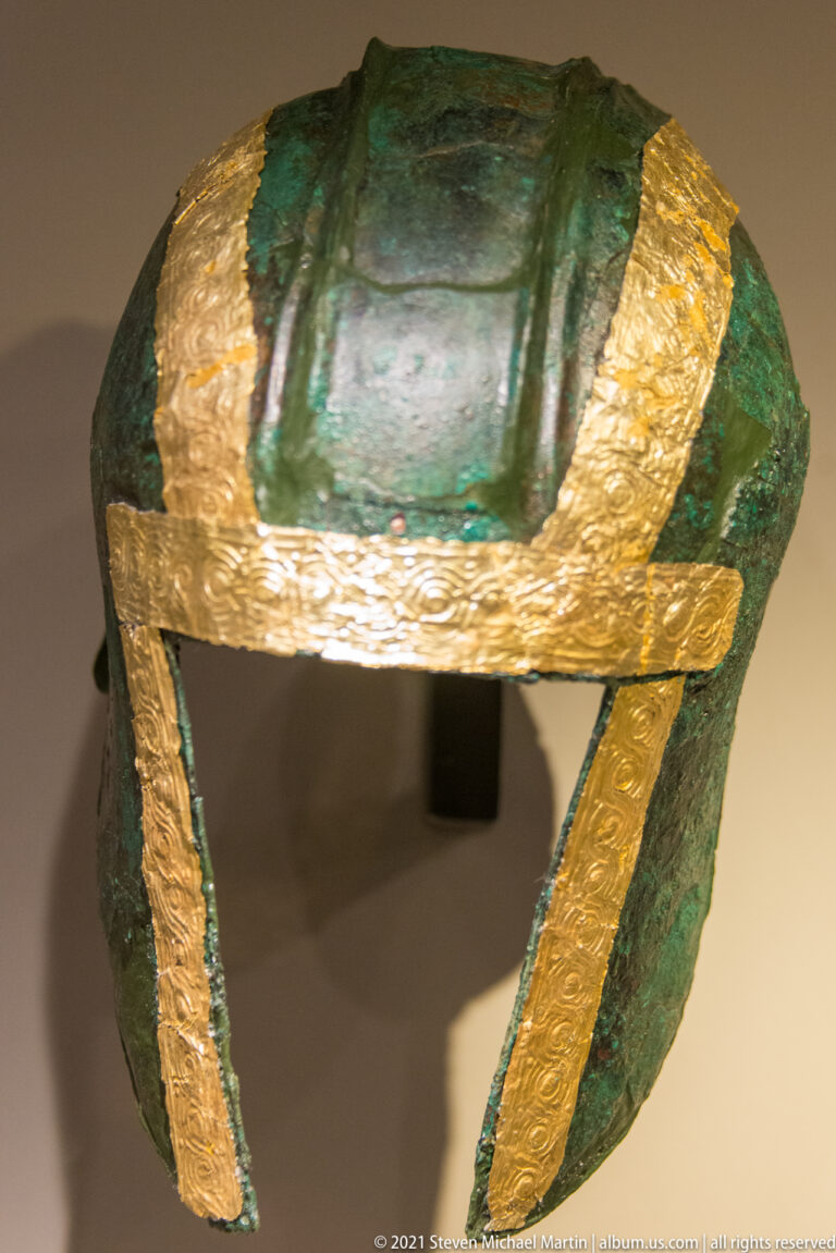 Green-patinated bronze helmet with broad gold strips bordering the top and sides of its open face.