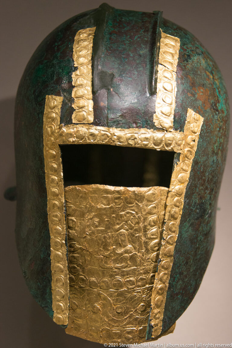 Rounded green-patinated bronze helmet with vertical embossed gold strips and a large wrinkled gold lower plate.