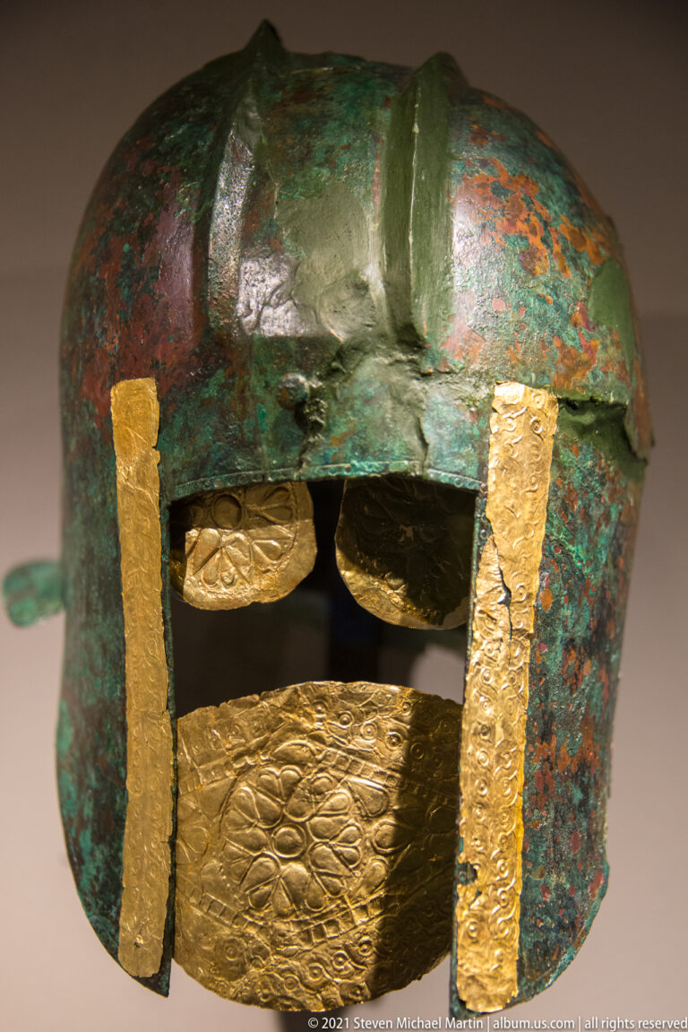 Bronze helmet with green and reddish patina, gold side strips, and several embossed plaques across the lower front.