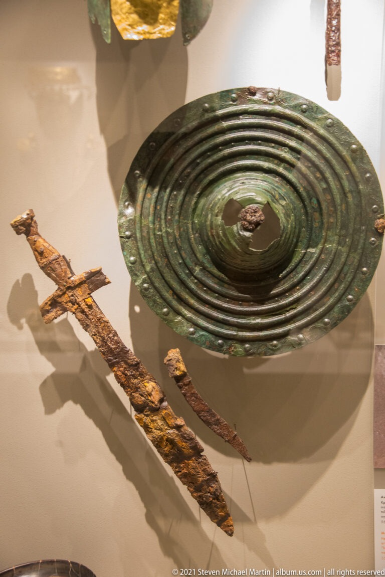 Round bronze shield with concentric rings displayed above several corroded iron blades or weapons.