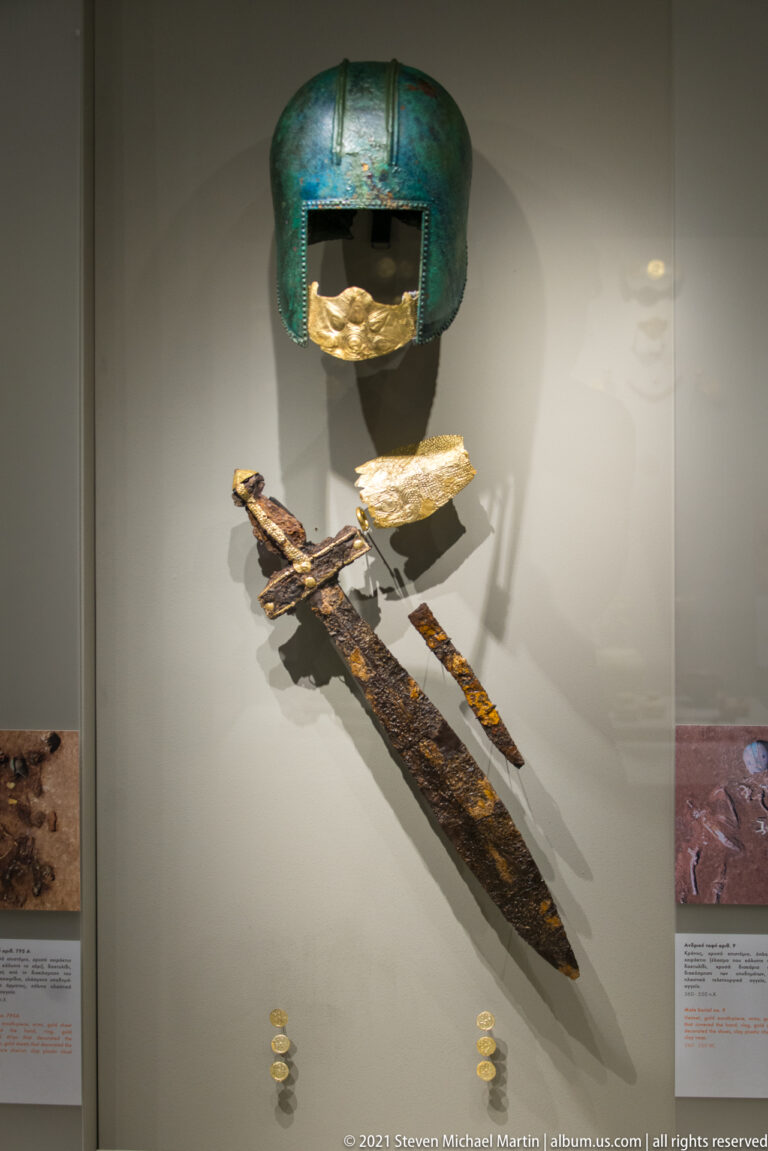 Bronze helmet above a corroded sword and dagger, with a gold fitting and small gold disks in a museum case.
