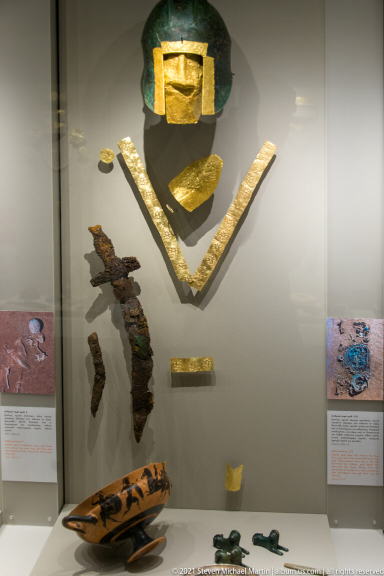 Helmet above V-shaped gold bands, corroded iron objects, a painted ceramic vessel, and small grave goods.