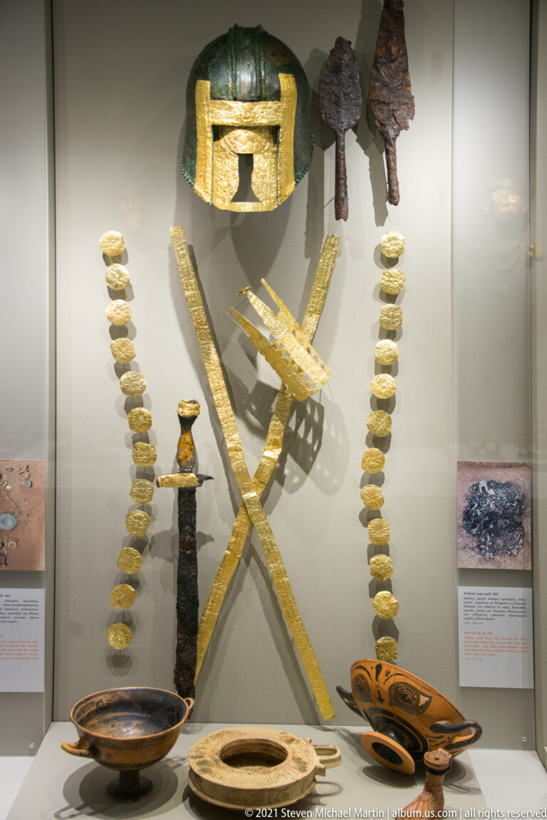 Bronze helmet above crossed gold bands, weapons, pale bead strands, and several ceramic vessels.