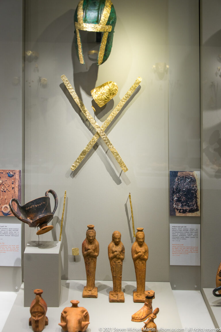 Bronze helmet and crossed gold bands above terracotta figurines and painted ceramic vessels in a museum case.
