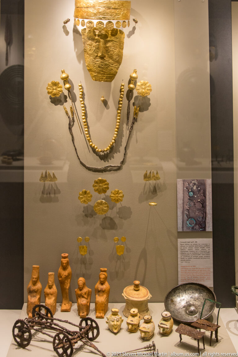 Gold ornaments and necklaces above terracotta figurines, miniature pottery, and metal objects in a museum display.