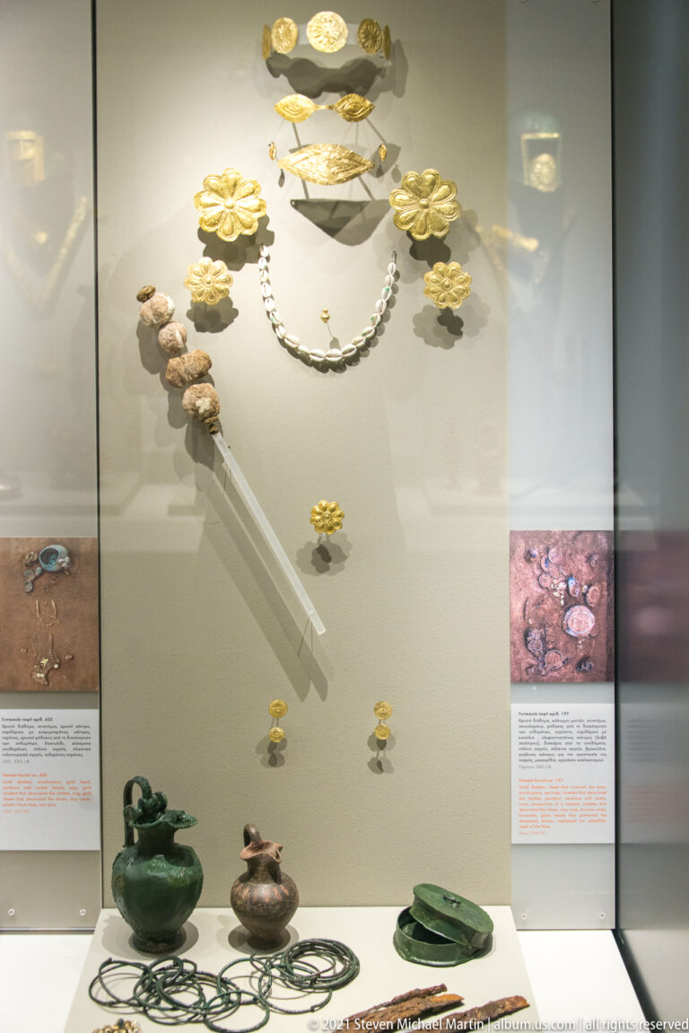 Gold rosettes, bands, necklaces, pins, bronze vessels, and other grave goods arranged in a museum case.