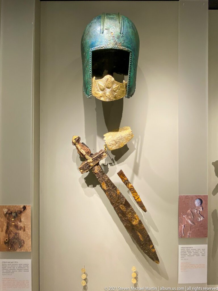 Green bronze helmet above gold fittings and a long corroded iron blade with surviving gold decoration.