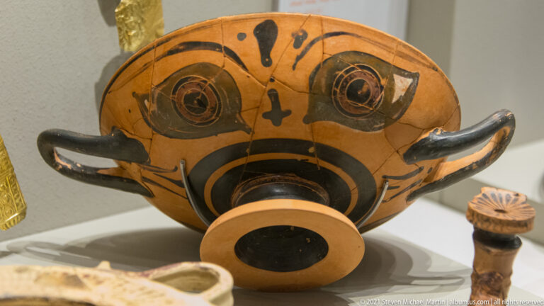 Two-handled orange ceramic vessel painted with large circular eyes and black bands forming a face-like design.