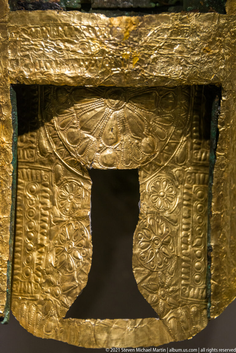 Close view of an embossed gold helmet panel with floral motifs, geometric borders, and a central opening.