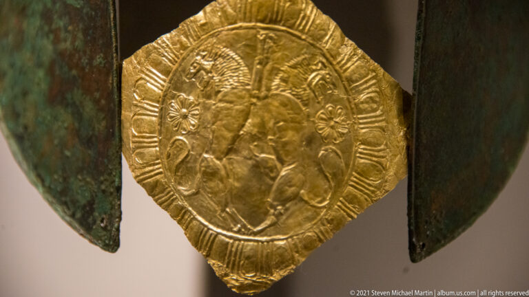 Embossed oval gold medallion with a worn multi-figure scene suspended between green-patinated bronze helmet sides.