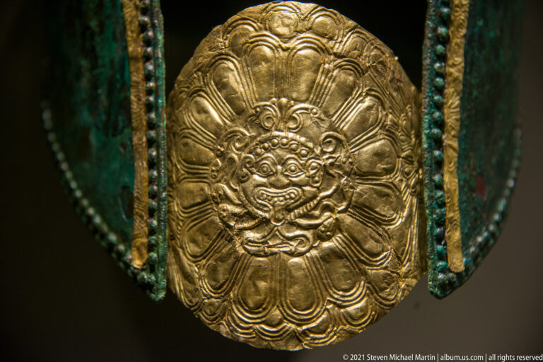 Embossed oval gold appliqué with a frontal mask-like face surrounded by layered petal forms.