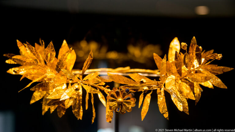 Side view of a gold wreath with dense pointed leaves and flower clusters projecting from a thin branch.