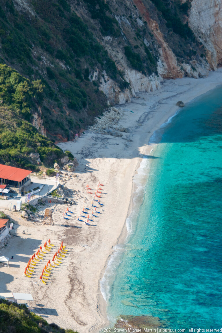 High view of Paralia Petani with rows of orange umbrellas, blue loungers, pale beach, turquoise sea, and steep cliffs.