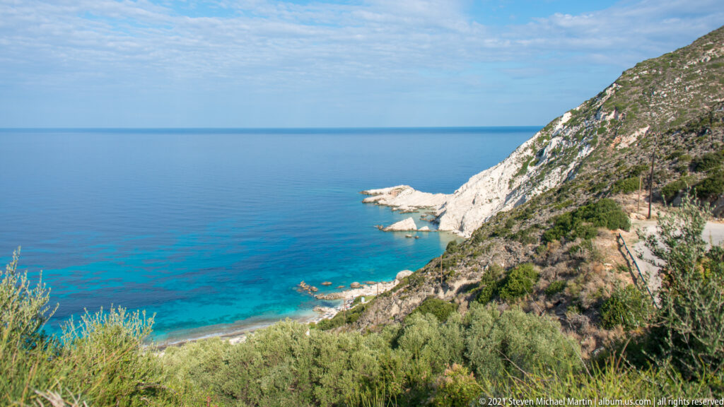 Steep vegetated slope descends to pale limestone rocks and turquoise shallows beside a calm blue sea.