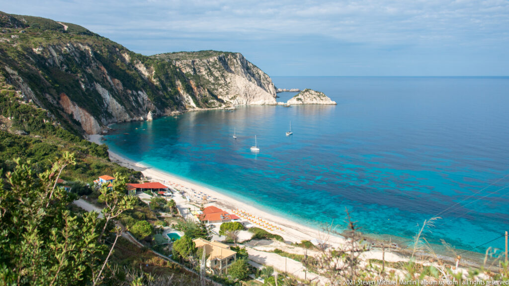 Elevated view across Paralia Petani, with a pale beach, turquoise bay, anchored sailboats, cliffs, and a rocky islet.