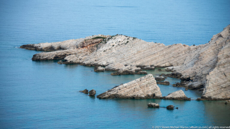 Layered pale limestone slabs tilt diagonally into calm blue seawater, with angular detached rocks along the shore.