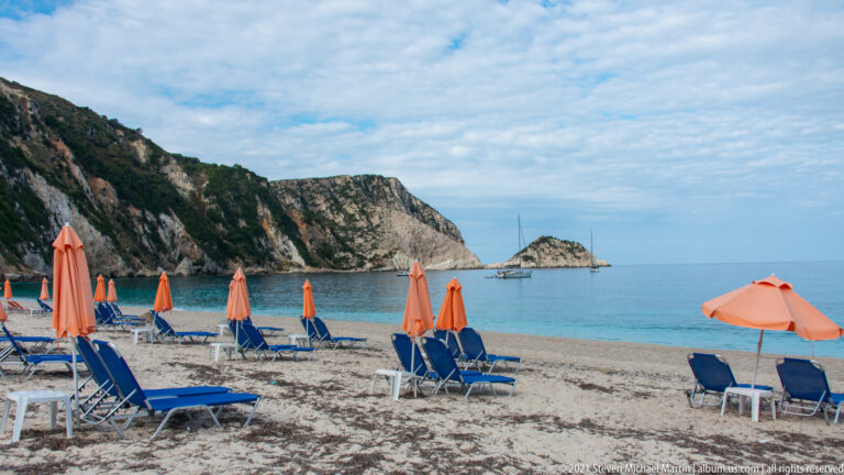 Blue loungers and orange umbrellas line the beach facing calm water, a sailboat, cliffs, and a small rocky islet.
