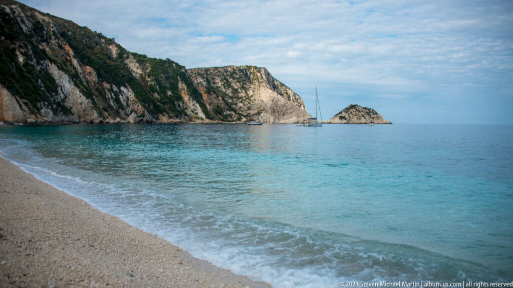 Pale pebble beach meets clear turquoise surf, with cliffs, a rocky islet, and anchored sailboats across the bay.