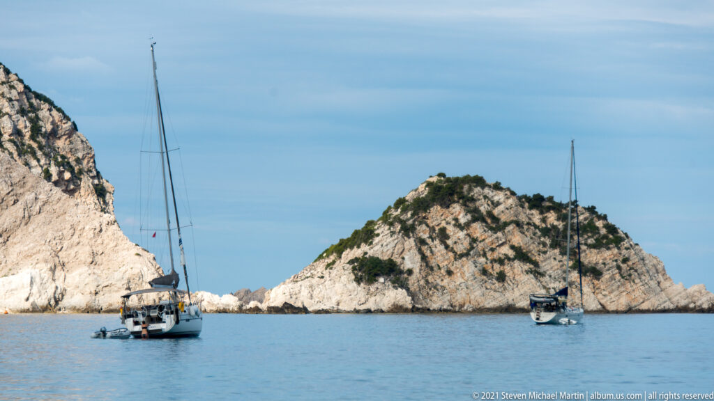 Two sailboats anchor in calm blue water on either side of a steep pale rocky islet near coastal cliffs.