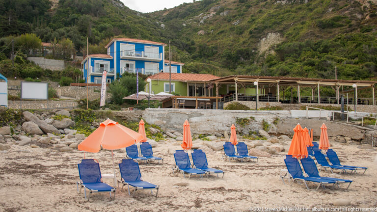 Orange umbrellas and blue loungers stand on a stony beach below open-air dining structures and hillside buildings.