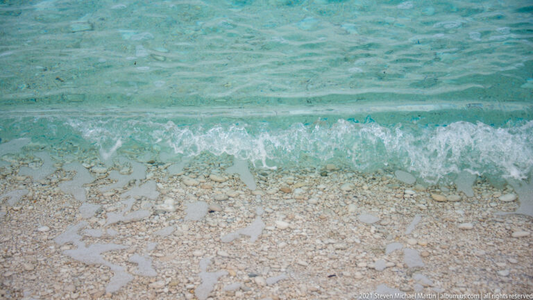 Small turquoise wave breaks over pale rounded pebbles visible through exceptionally clear shallow water.