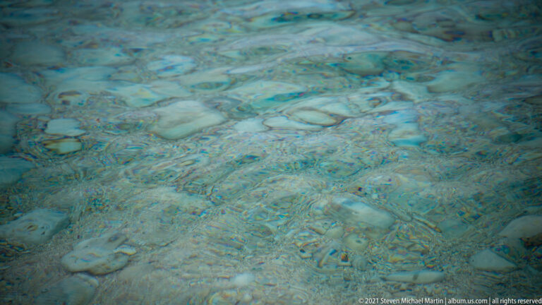 Rounded pale pebbles show through clear blue-green seawater, with surface ripples distorting their shapes.