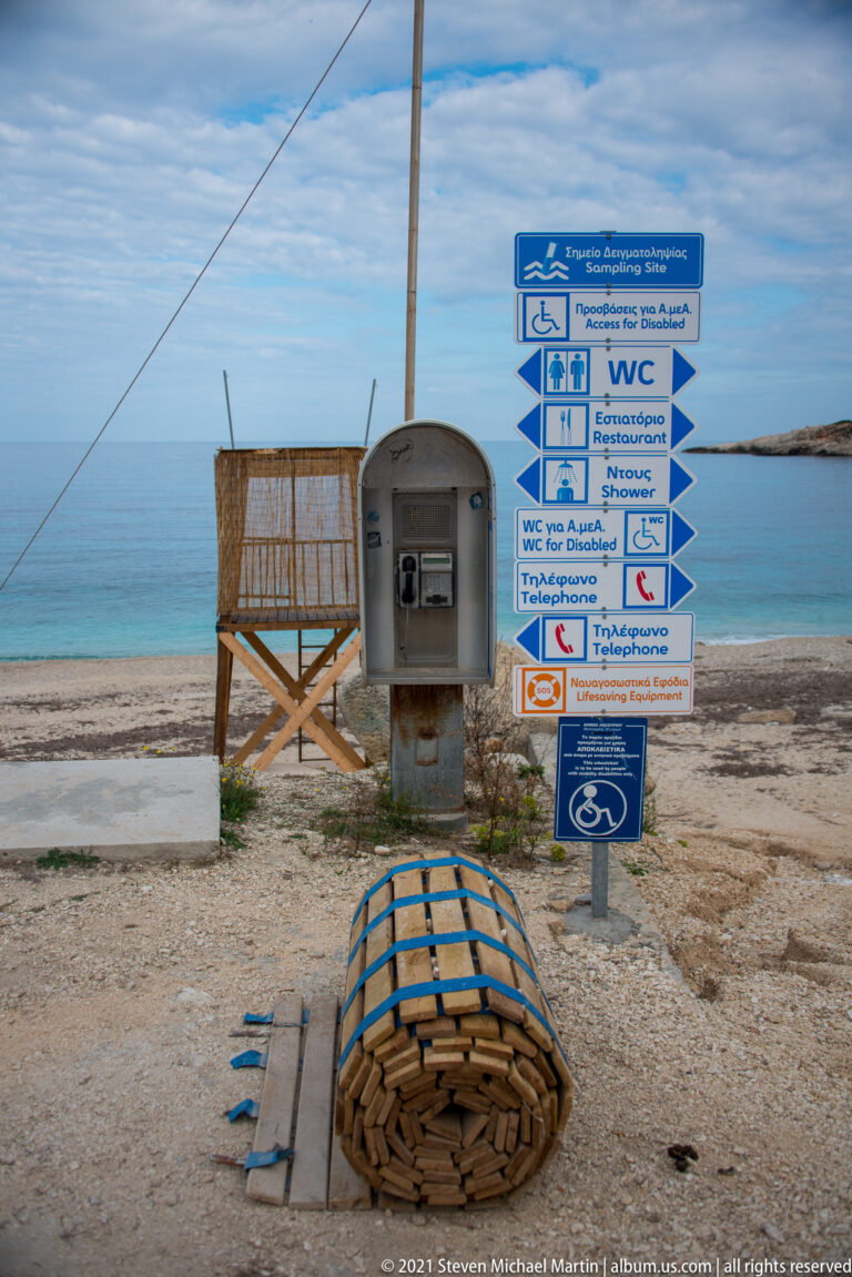 Blue-and-white beach service signs stand beside a public telephone, with stacked beach equipment and the sea behind.