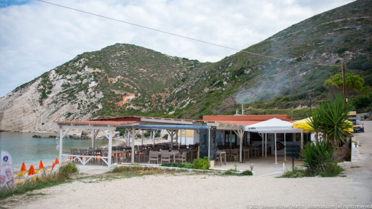 Open-air restaurant with shaded tables and awnings stands between the beach and a steep vegetated hillside.