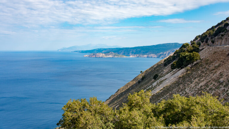 Steep rocky slope drops to blue sea, with shrubs in the foreground and layered coastal cliffs extending into the distance.