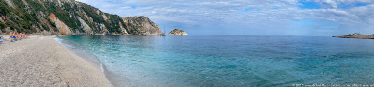 Wide beach-level view of clear turquoise water, pale shore, a towering cliff, and a small offshore rock.