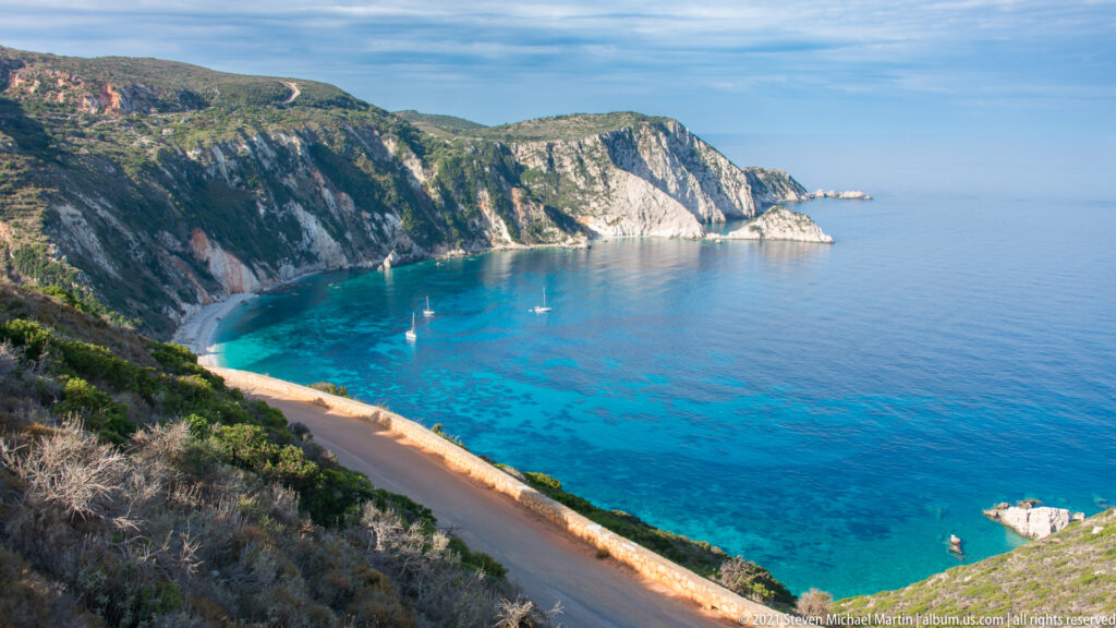 Road curves above a turquoise bay with anchored sailboats, a narrow beach, and pale limestone cliffs.