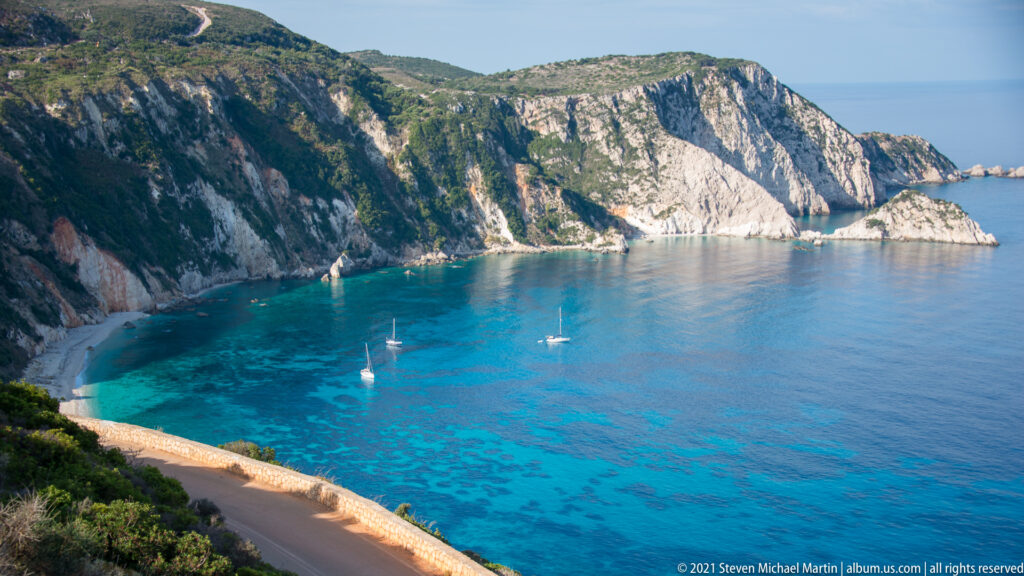 Three sailboats anchor in turquoise water below high pale cliffs, with part of Paralia Petani visible at left.
