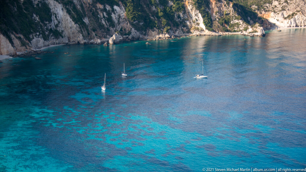Several sailboats anchor in clear turquoise-blue water below pale limestone cliffs enclosing a coastal bay.