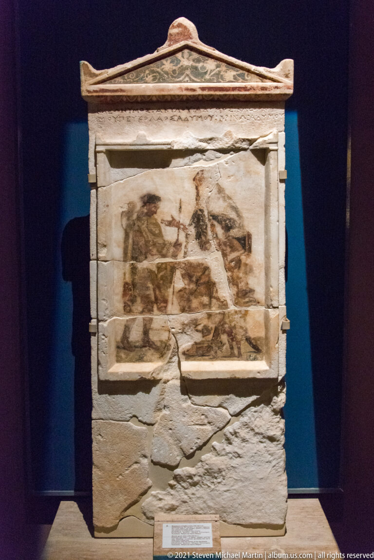Tall pedimented stone grave monument with a fragmentary painted panel of human figures above a cracked lower section.