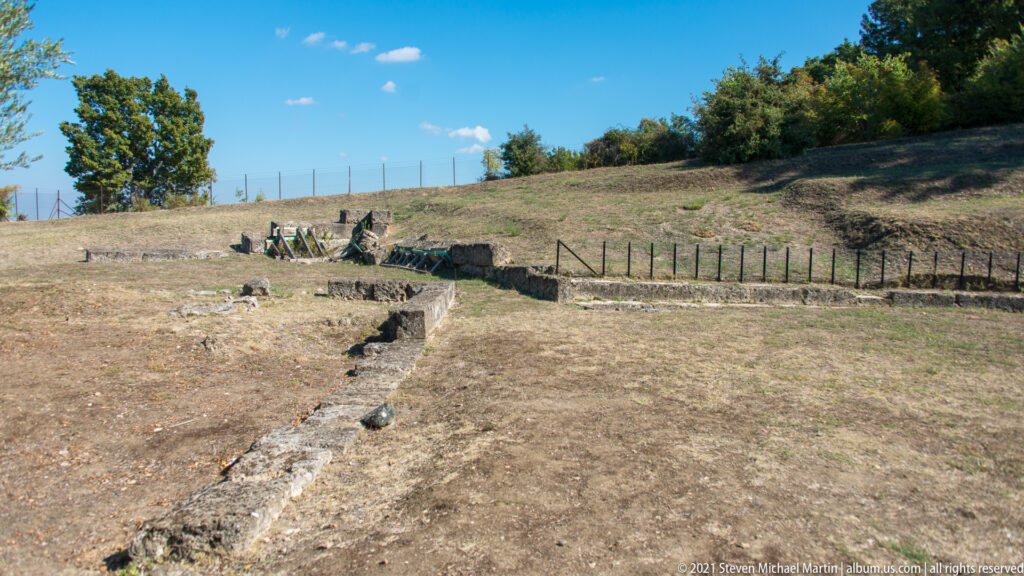 Low ancient stone foundations and retaining walls on a dry grassy slope, bordered by railings and trees.
