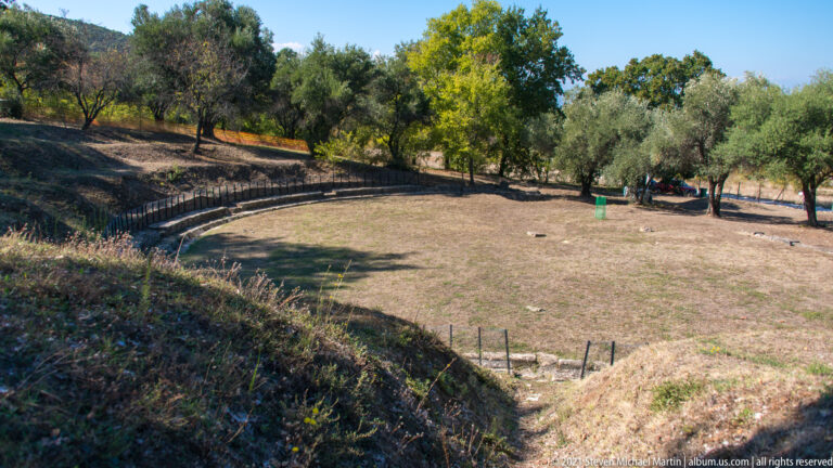 Curving rows of ancient stone seating surround a broad grassy theater area bordered by trees and protective railings.