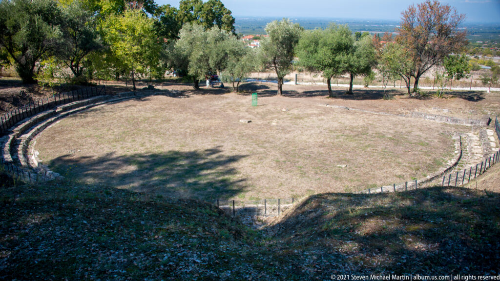 Elevated view of semicircular stone theater seating around a grassy center, with trees and a distant plain beyond.