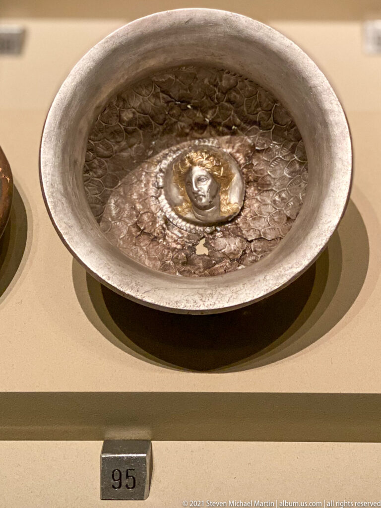 Shallow silver bowl with a broad rim, textured interior, and raised circular human-face medallion at the center.