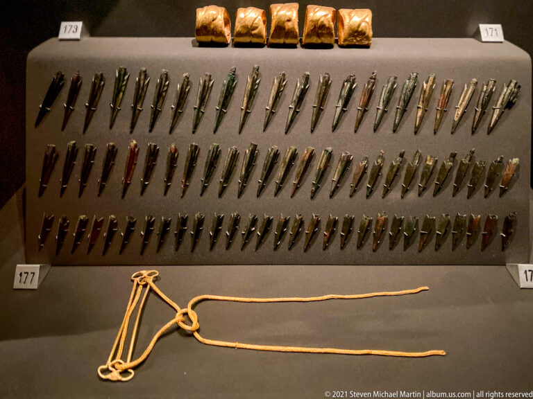 Rows of narrow metal arrowheads and small weapon fittings beneath several gold-covered pieces, with a long cord displayed below.