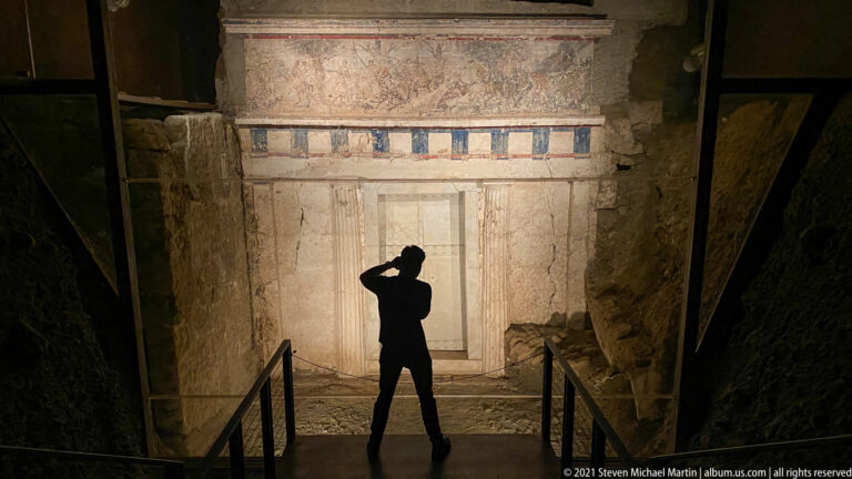 Silhouetted visitor facing an illuminated Macedonian tomb façade with a central doorway and blue-and-white painted frieze.